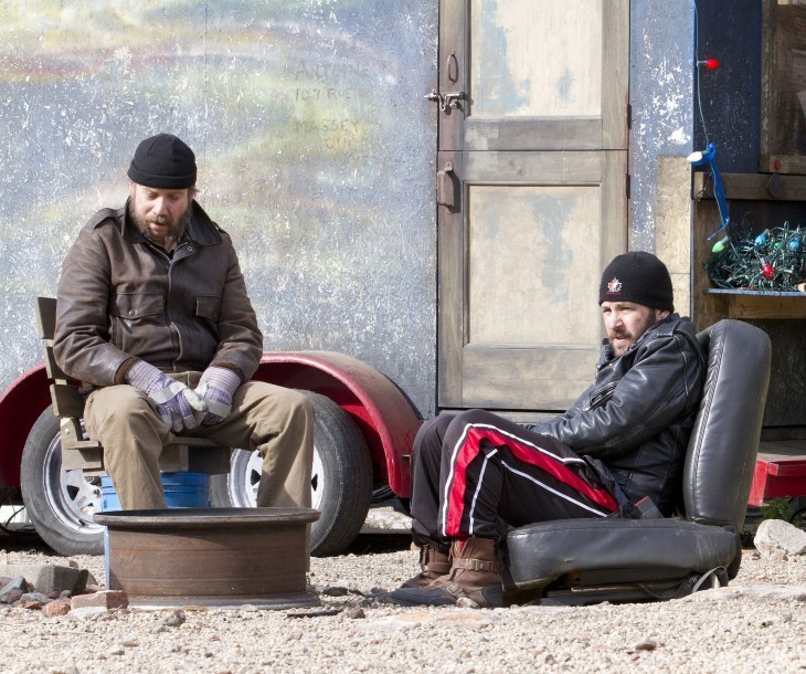Photo of Paul Giamatti and Paul Rudd, slumped over in a dirt lot with a trailer, looking cold and glum