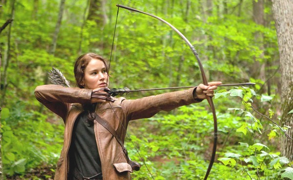 Photo of Katniss Everdeen, a young woman, drawing a bow and scowling in the forest