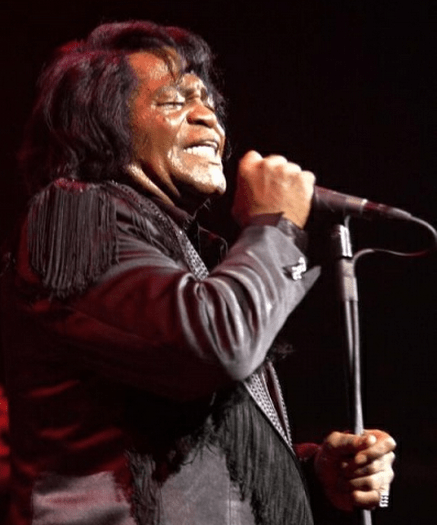 A photo of James Brown, lookin' older, crooning into a microphone gripped in both hands