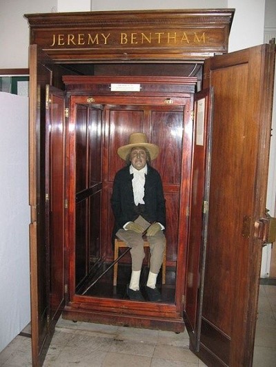 A photo of the body of Jeremy Bentham, looking like a Madame Tussaud's wax figure, sitting up in a glass-fronted cabinet