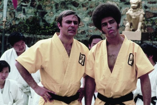 Two men in karate costumes with black belts stand side by side, looking grim