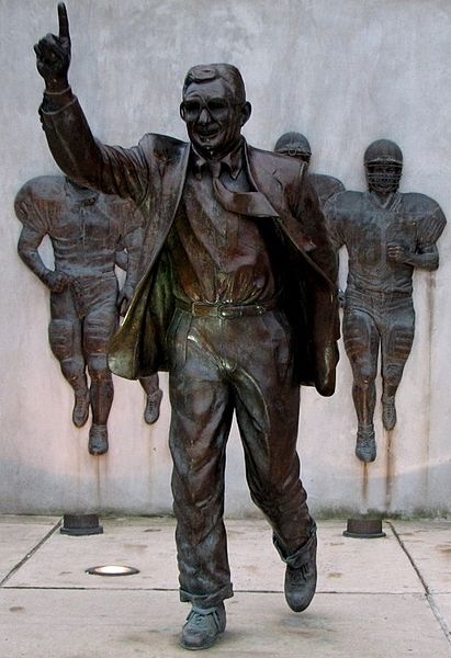 Photo of a bronze statue of Joe Paterno, running with one finger held high in front of bronze football players
