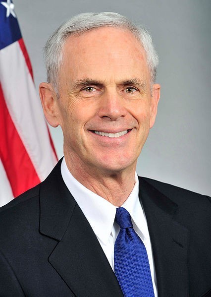 A photo of John Bryson as Secretary of Commerce, with a blue suit and tie in front of a flag