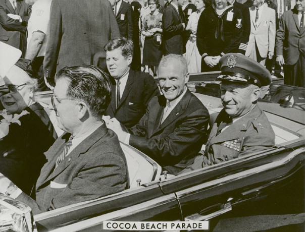 Photo of John Glenn in the back seat of a convertible limo, between John Kennedy and a general