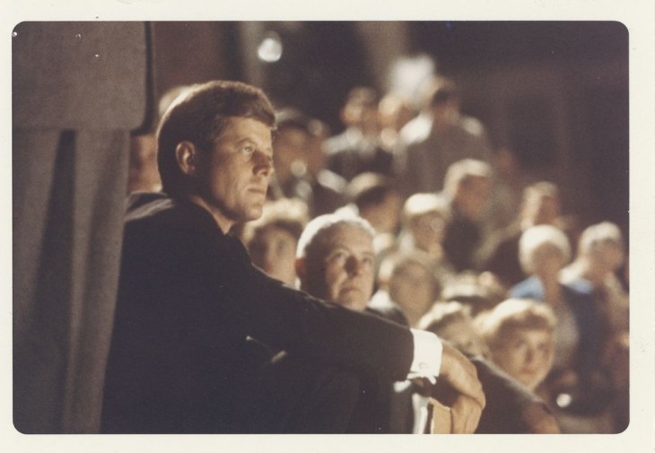 Photo of John F. Kennedy sitting and listening on steps among a crowd
