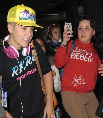 Photo of Justin Bieber walking through an airport, while a teenaged girl in a red Bieber shirt takes a photo intently