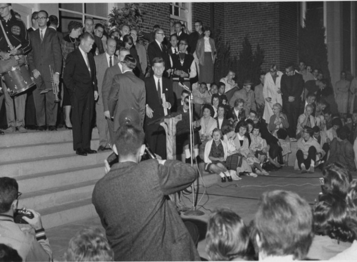 Black and white photo of John F. Kennedy on steps, speaking, surrounded by students