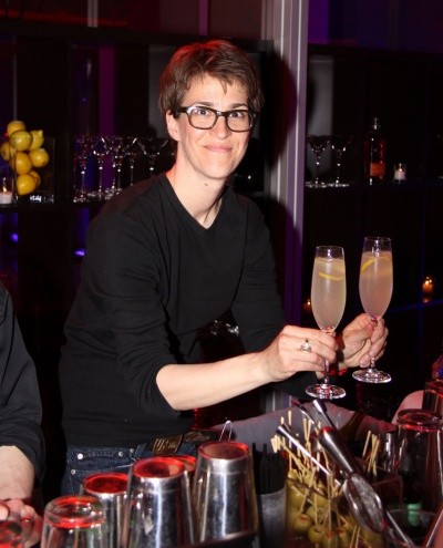 A photo of Rachel Maddow holding two long-stemmed glasses with pink liquid and lemon twists