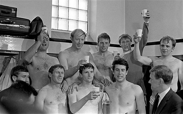 A photo of two rows of skinny shirtless pasty-skinned 30ish guys raising paper cups of champagne in an old locker room
