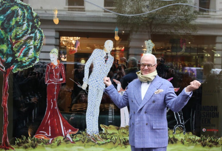 A photo of Manolo Blahnik standing in a natty blue suit in front of a store fashion display window