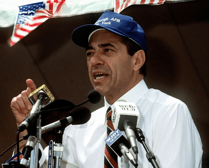 Photo of Mario Cuomo, in shirtsleeves and tie and a baseball cap, speaking behind a podium of microphones