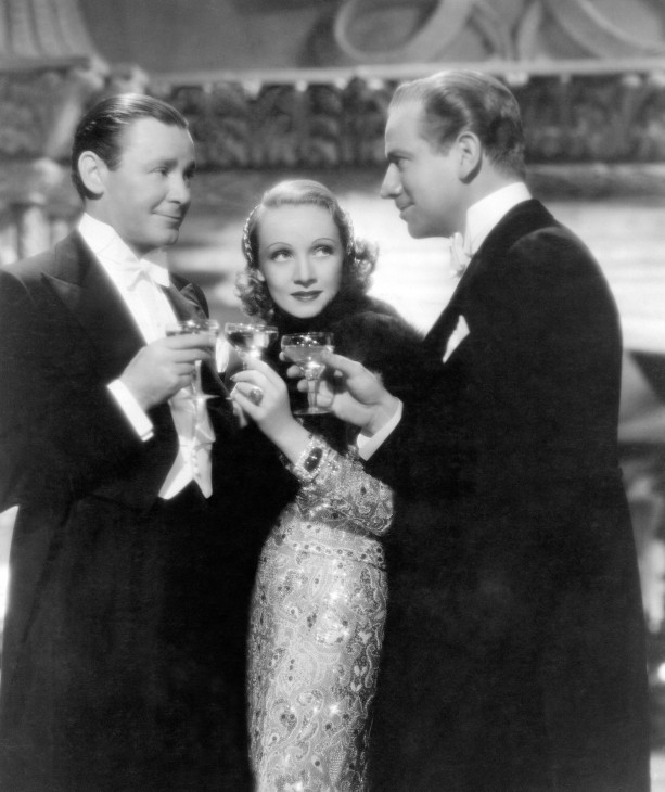 Marlene Dietrich is glam and beautiful in a slinky embroidered gown. She stands between two slicked-back men in tuxedoes who are staring intently at each other. All three are holding cocktail glasses in some kind of fancy mansion or hotel lobby.
