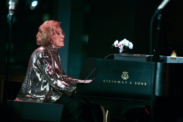 A photo of Marian McPartland in a silver jacket, sitting at a Steinway piano