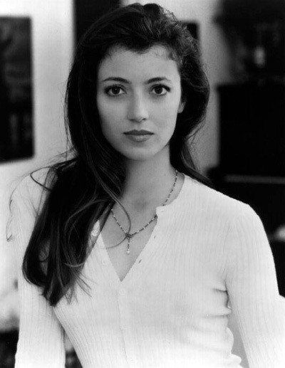 Mia Sara seen from the waist up in a white sweater, with long black hair tossed down over her left shoulder. Her eyes are round and dark, her lips full.
