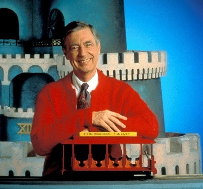 A photo of Mister Rogers standing with the Neighborhood Trolley in a red cardigan sweater