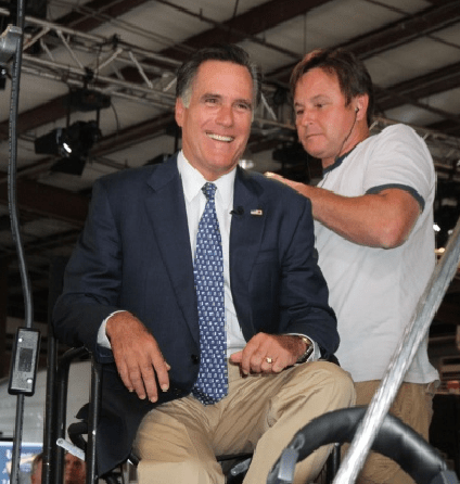 Mitt Romney smiles onstage as a sound technician fixes wires to his back