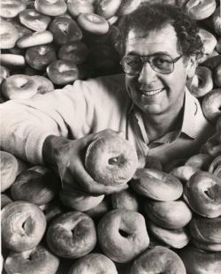 A photo of Murray Lender in a pile of bagels, smiling as he holds a bagel up to the camera