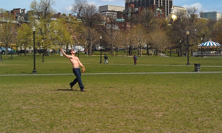 Photo of a shirtless man tossing a baseball to a friend on the green grass of Boston Common