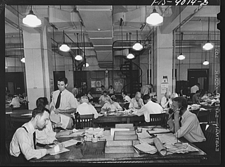 Photo of newsmen in suspenders and ties, working at a series of wooden desks