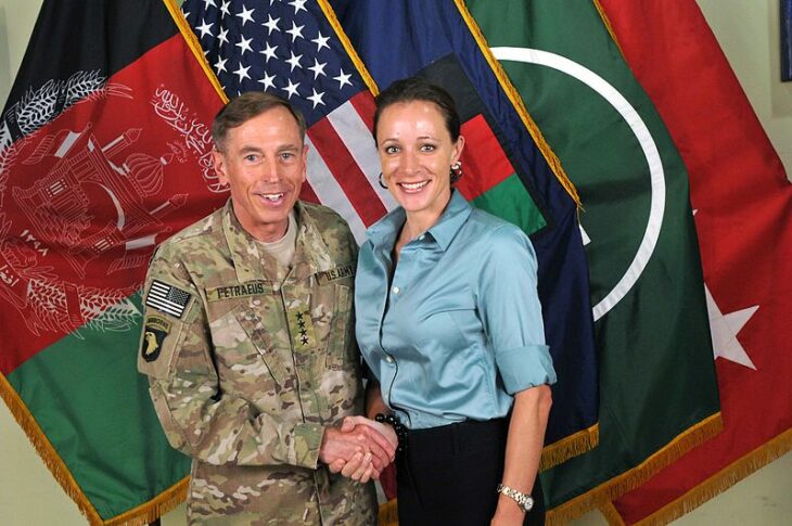 A general in combat fatigues shakes hands with a woman in a blue shirt in front of flags