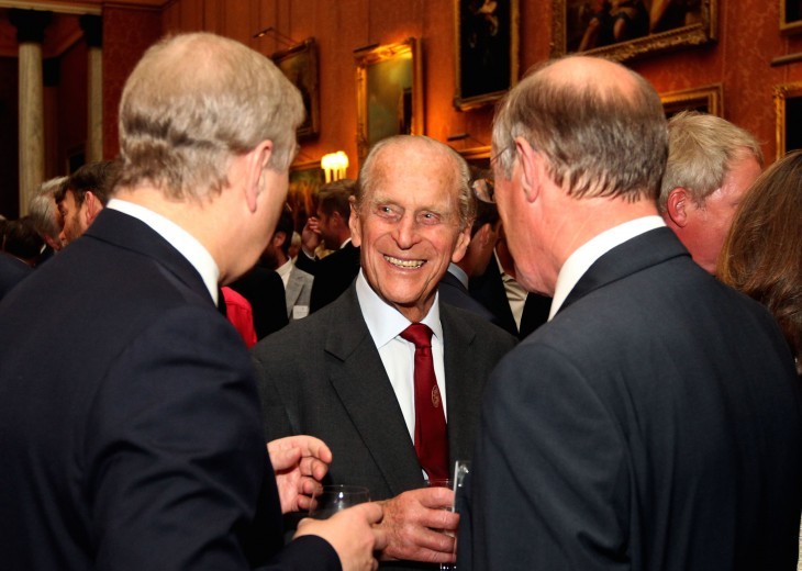 Prince Philip, drink in hand, smiles at two other gents as they joke at a formal club of some kind