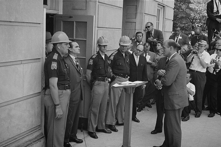 A photo of George Wallace standing in a doorway, surrounded by his own police, facing a balding man with his arms crossed