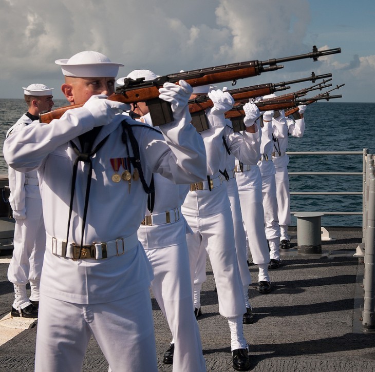 Sailors dressed in white fire their guns over the water