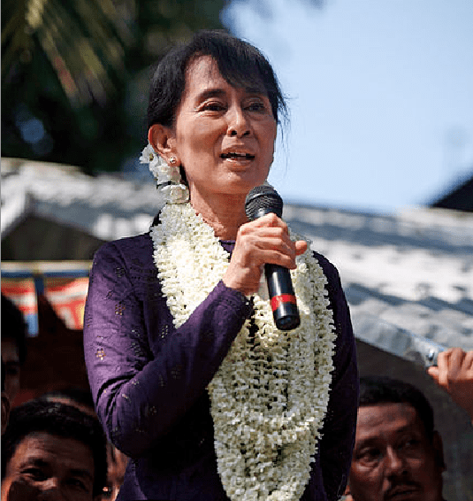 Photo of Aung San Suu Kyi, draped in leis (or something like them) and holding a big microphone, talking to smiling supporters