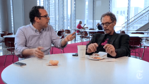 Two men in casual clothes sit at a cafeteria table in a room with huge windows and great light