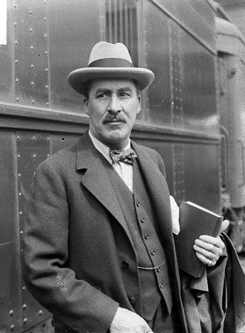 Photo of Howard Carter in a suit, standing before a train car and holding a book