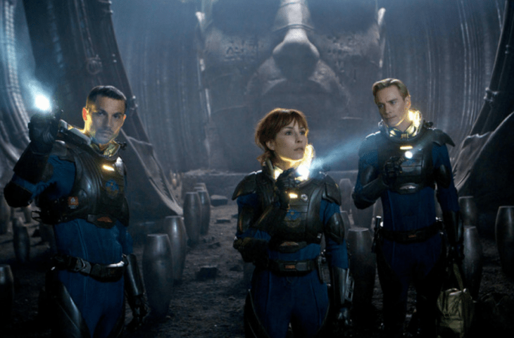 A photo of Noomi Rapace in 'Prometheus,' shining a flashlight in an old cave with 2 other astronauts, a huge stone head behind