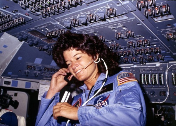 A photo of Sally Ride in a blue flight suit, floating in zero gravity, smiling and talking on a headset