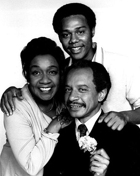 Photo of Sherman Helmsley posing with his co-stars in a dark suit holding a carnation