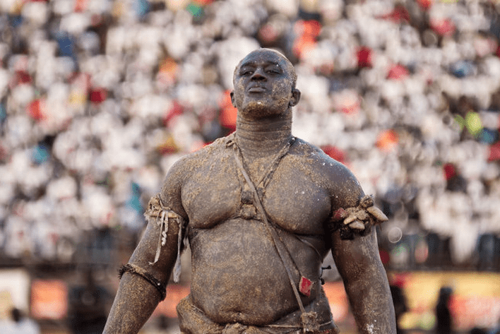 Photo of a muscular Senegal man, caked in dirt, with little sachet bags tied to his arms and a talisman across his shoulder