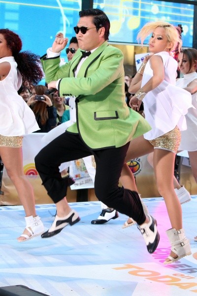 Photo of Psy on stage, galloping with his arms crossed as lady backup dancers do the same