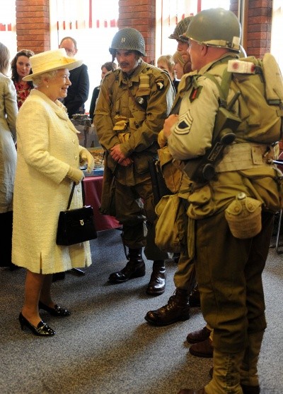Queen Elizabeth photo in bright yellow dress and hat, plus pumps and purse, talking to men dressed as WWII soldiers