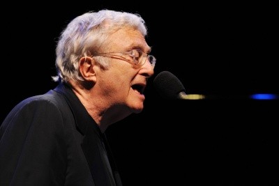 Photo of Randy Newman singing at a piano, mouth open to the microphone