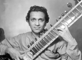 A photo of Ravi Shankar looking boldly at the camera with a sitar angled up in his lap
