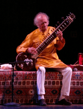 A photo of Ravi Shankar playing the sitar in a gold silk shirt on a big bench