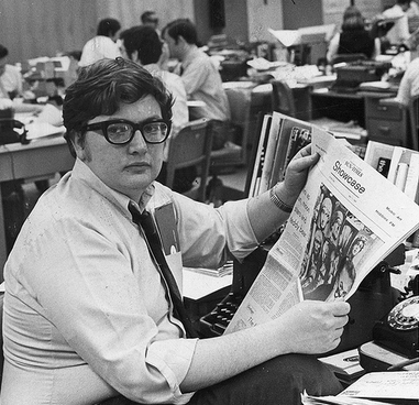 Photo of Roger Ebert, young with black horn-rim glasses, sitting in a newsroom