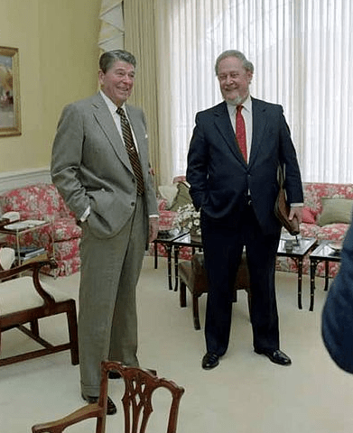 Photo of two older men, smiling, as they stand together in a nice carpeted office