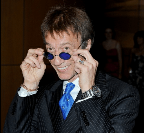 Photo of Robin Gibb in blue suit and tie, tipping his blue-tinted spectacles forward to show his eyes as he smiles for photogs