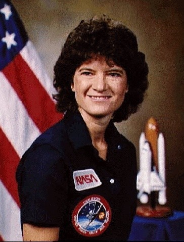 Sally Ride poses in a blue polo shirt in front of a flag and the NASA logo