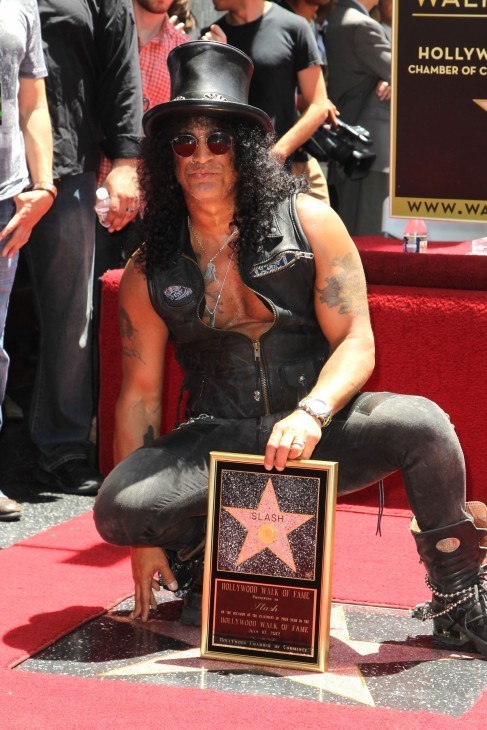 Photo of Slash in his top hat and leather vest and very long black hair, squatting behind his golden star on the sidewalk