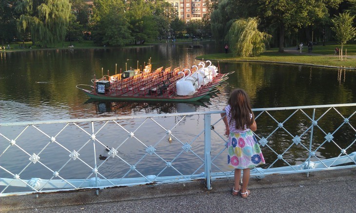 Photo of a little girl looking over a footbridge railing at swan boats tied up in the middle of a pond