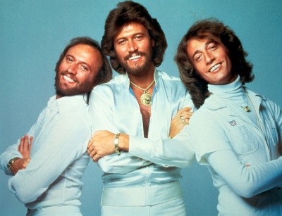 Photo of the Bee Gees with long hair and light blue jumpsuits, leaning in together and smiling