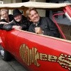Three middle-aged men smile cheerfully as they lean out the windows of an unusually long red convertible with the MONKEES guitar logo on the side