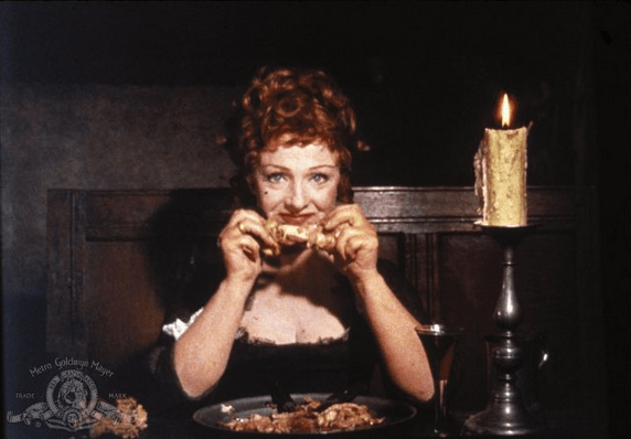 A photo of Joyce Redman, red-haired and 40ish, in a buxom dress and smiling as she holds a chicken leg near her mouth