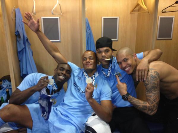 Four youngish muscular dark-skinned men smiling and crowding into a photo together in a modern wood-paneled locker room