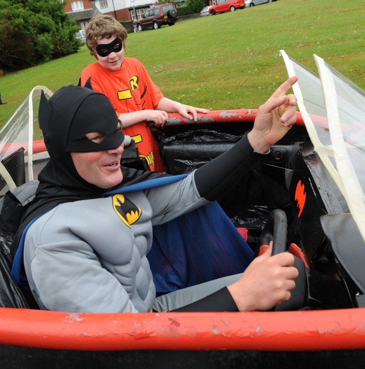 Batman costume man behind the wheel of a Batmobile, grinning